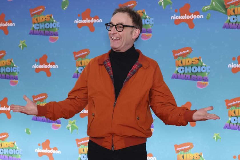 Tom Kenny in burnt orange jacket and black turtleneck before multicolor backdrop at the 2023 Nickelodeon Kids' Choice Awards