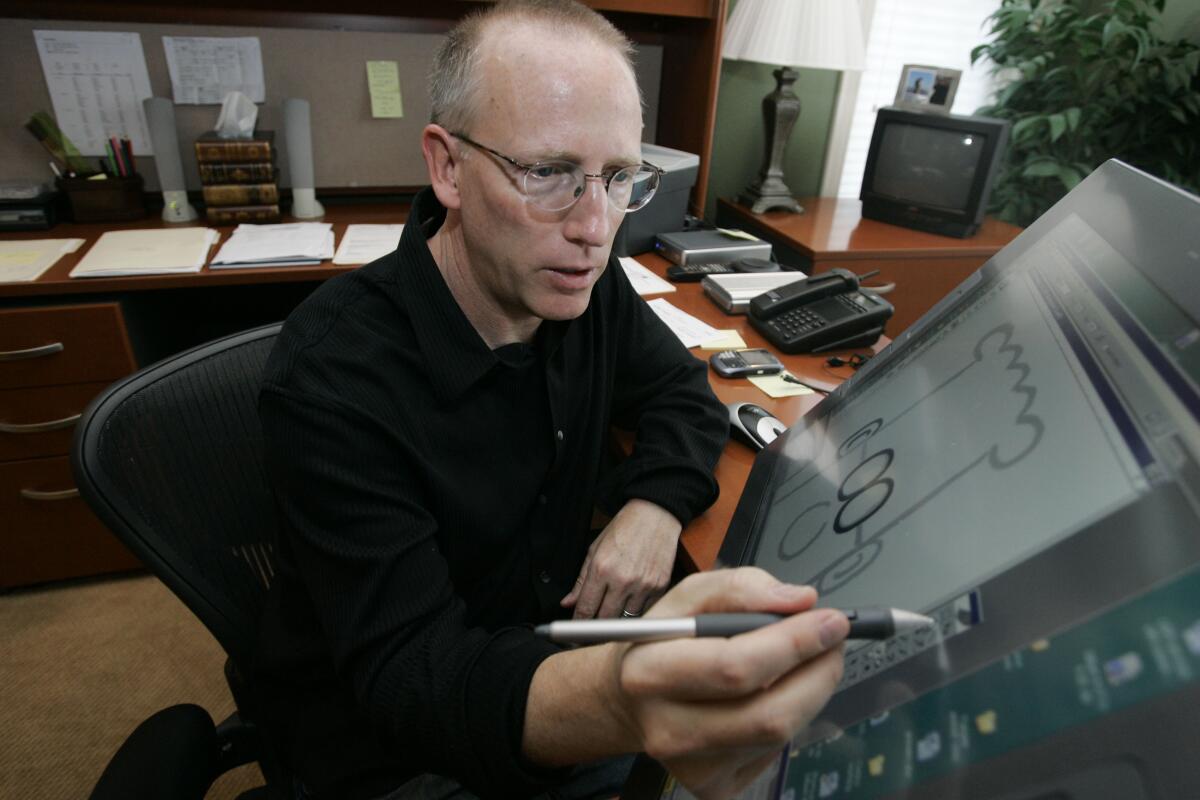 A balding man in glasses draws a comic at an art table in his studio