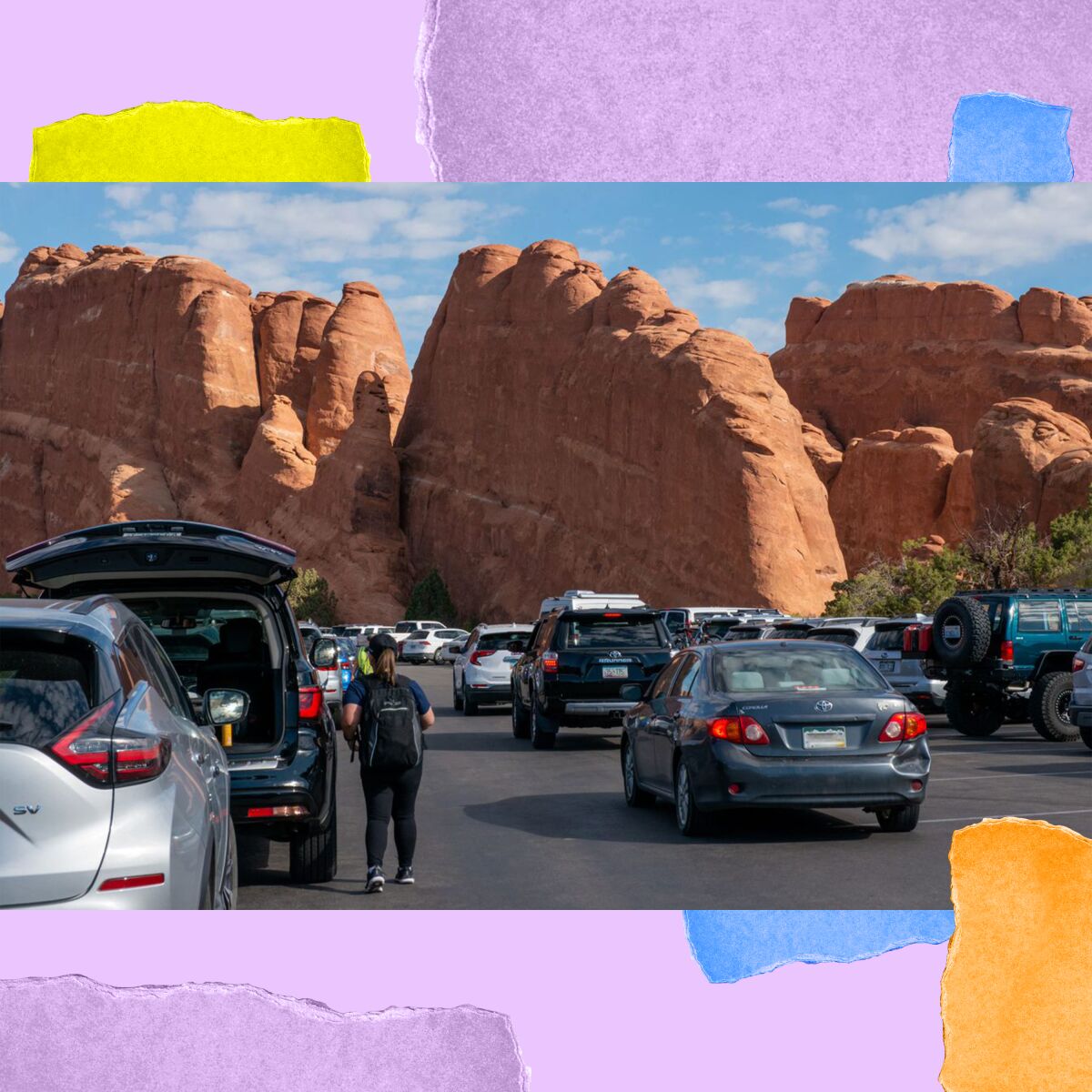 Cars are lined up in a parking area next to towering red rock boulders.