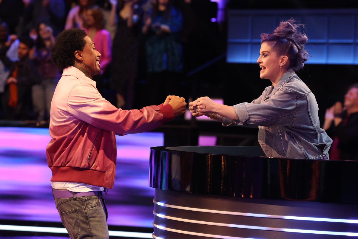 Nick Cannon shares a fist bump with Kelly Osbourne on a colorful game-show set.