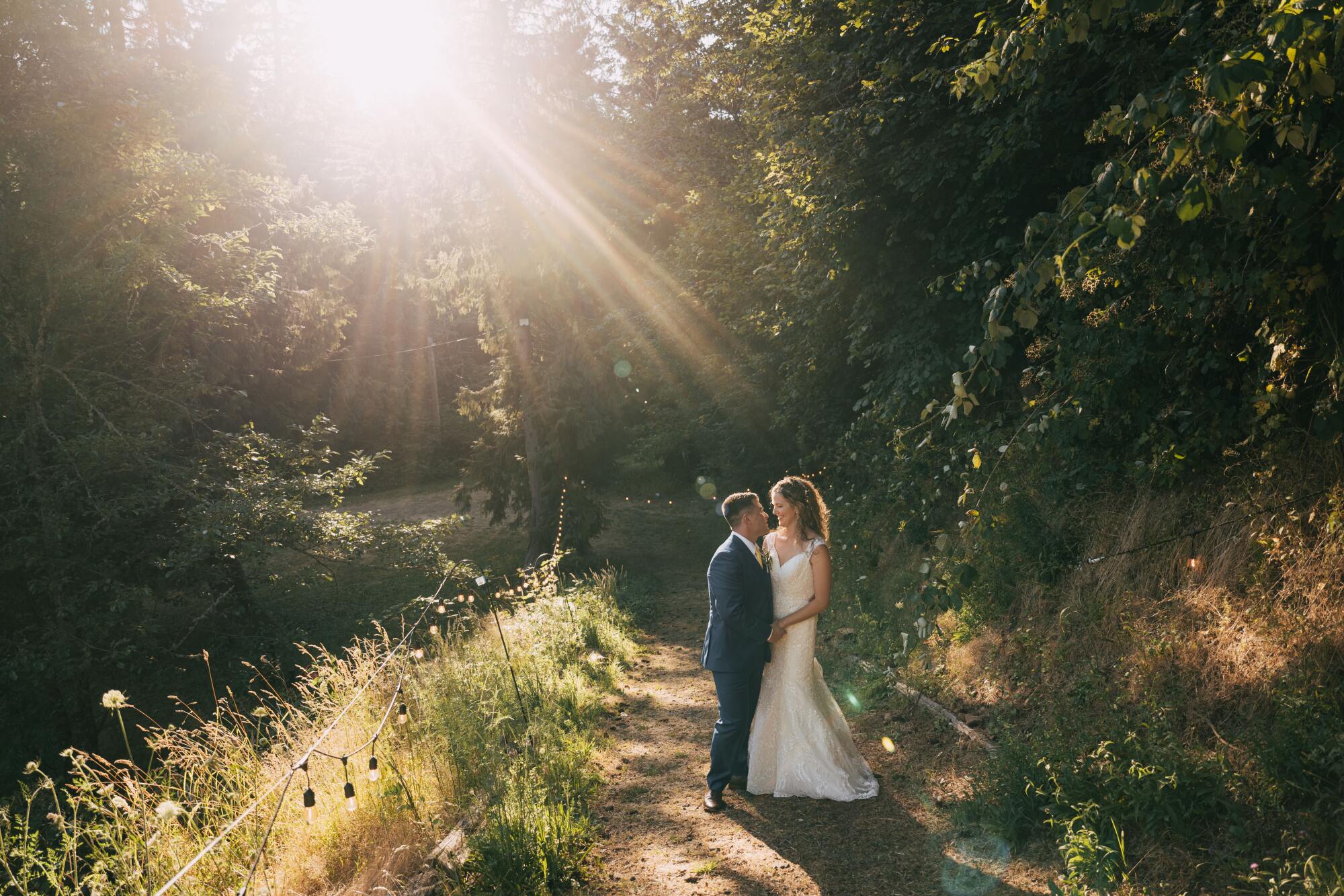 A bride and groom stand outdoors under rays of sunlight