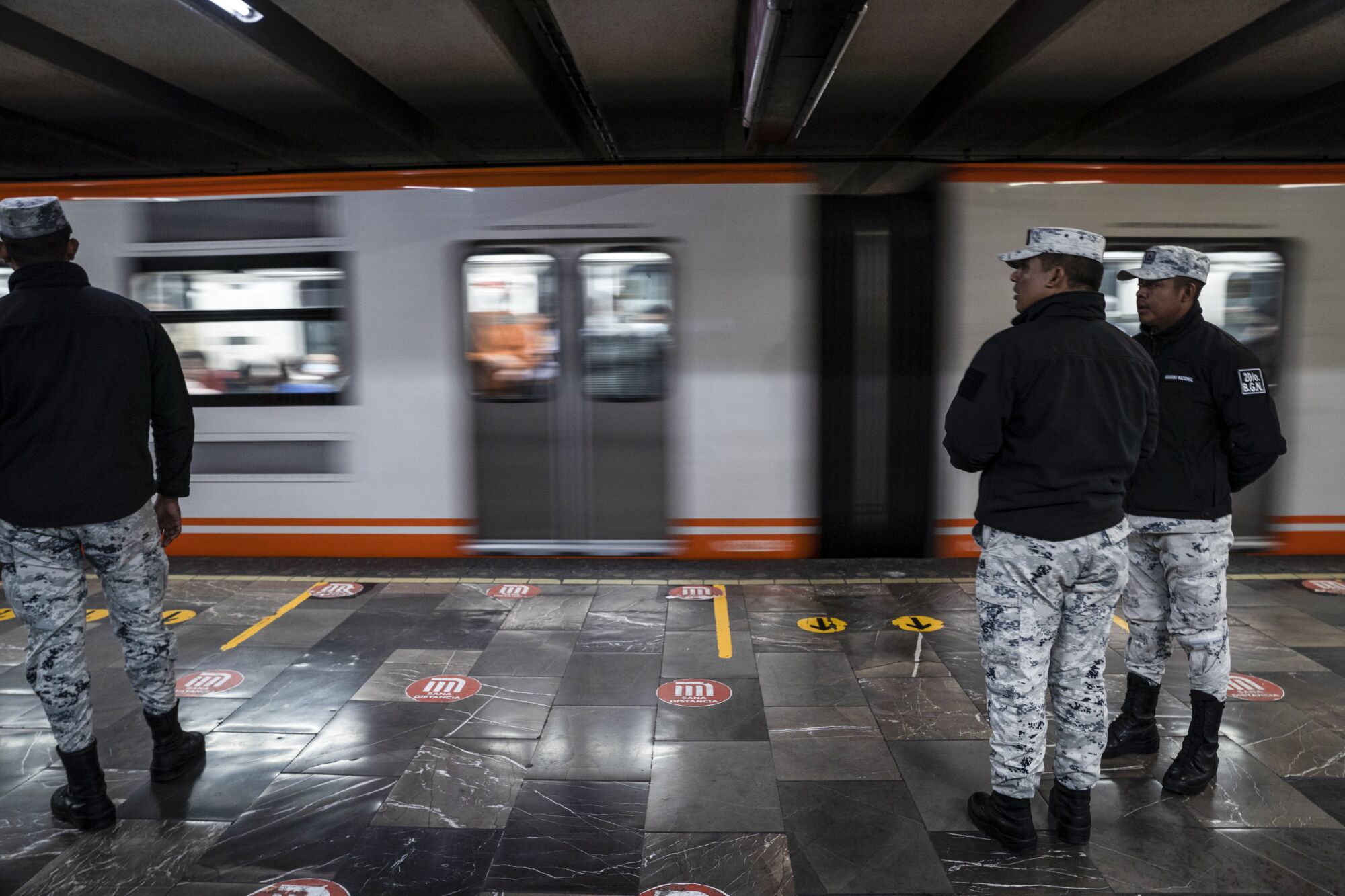 Bir metro treni hızla geçerken aynı kıyafetlere sahip üç kişi bir platformda duruyor.