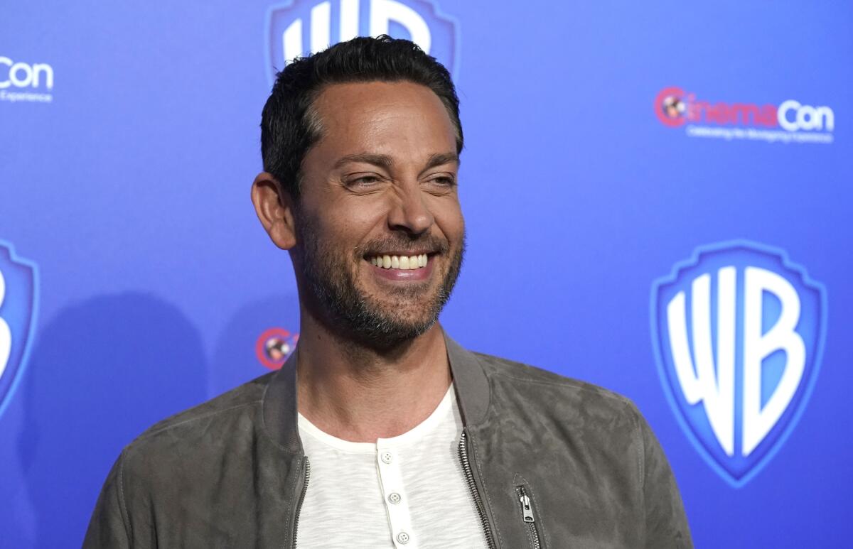 A man with short brown hair wearing a gray jacket and smiling against a blue background