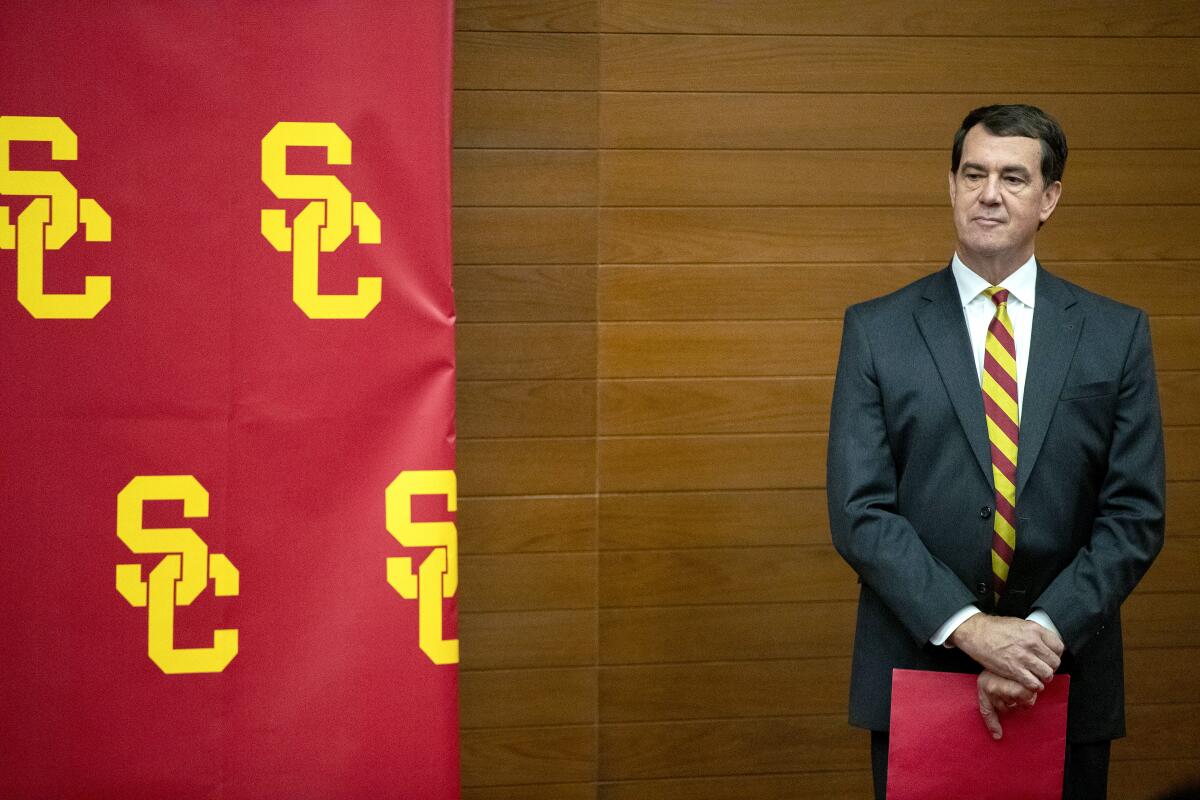 Mike Bohn holds a red folder on a stage with a red and yellow backdrop decorated with USC logos at left