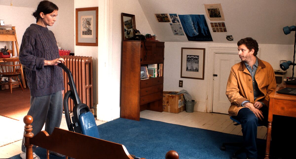 A woman vacuums while a man sits at a desk.