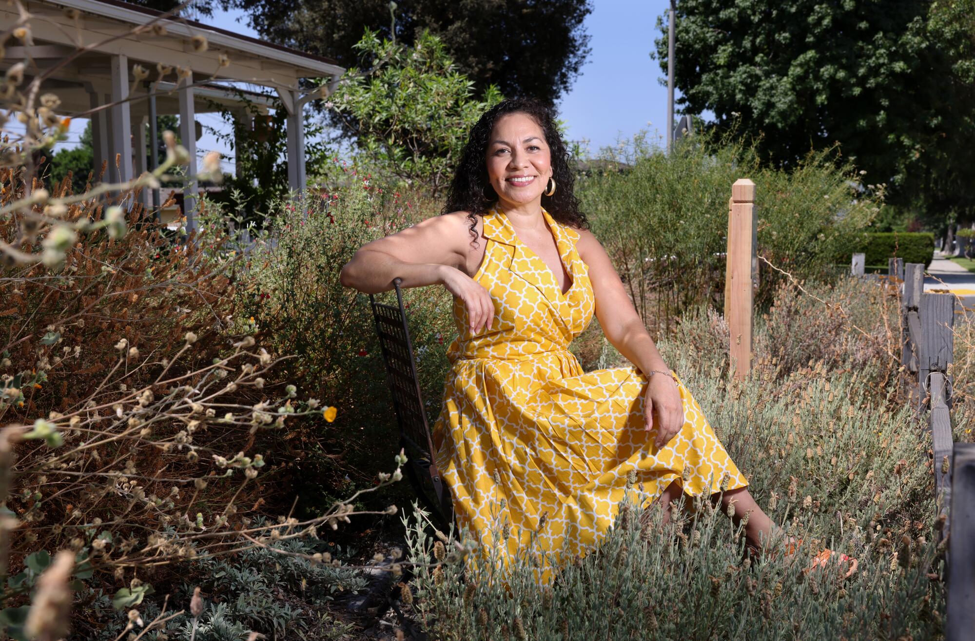 Aurora Anaya in a yellow sundress sits in her native plant garden, which is dull greens and browns during summer dormancy.