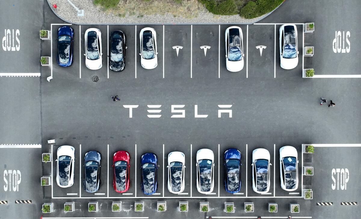 Cars sit in a parking lot with the word "Tesla" painted on the pavement in a bird's-eye view