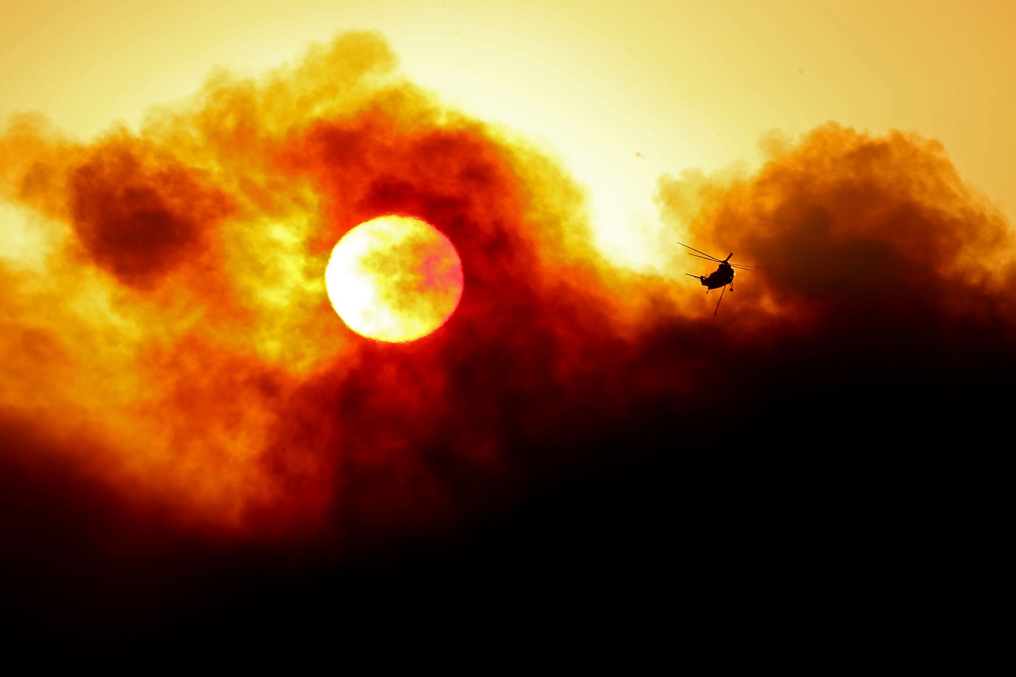 The sun is partially obscured by smoke as a firefighting helicopter prepares to make a water drop.