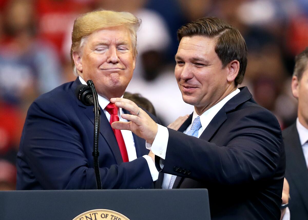 Donald Trump, at left, rests his right hand on the shoulder of Ron DeSantis, who is reaching for a microphone at a lectern. 