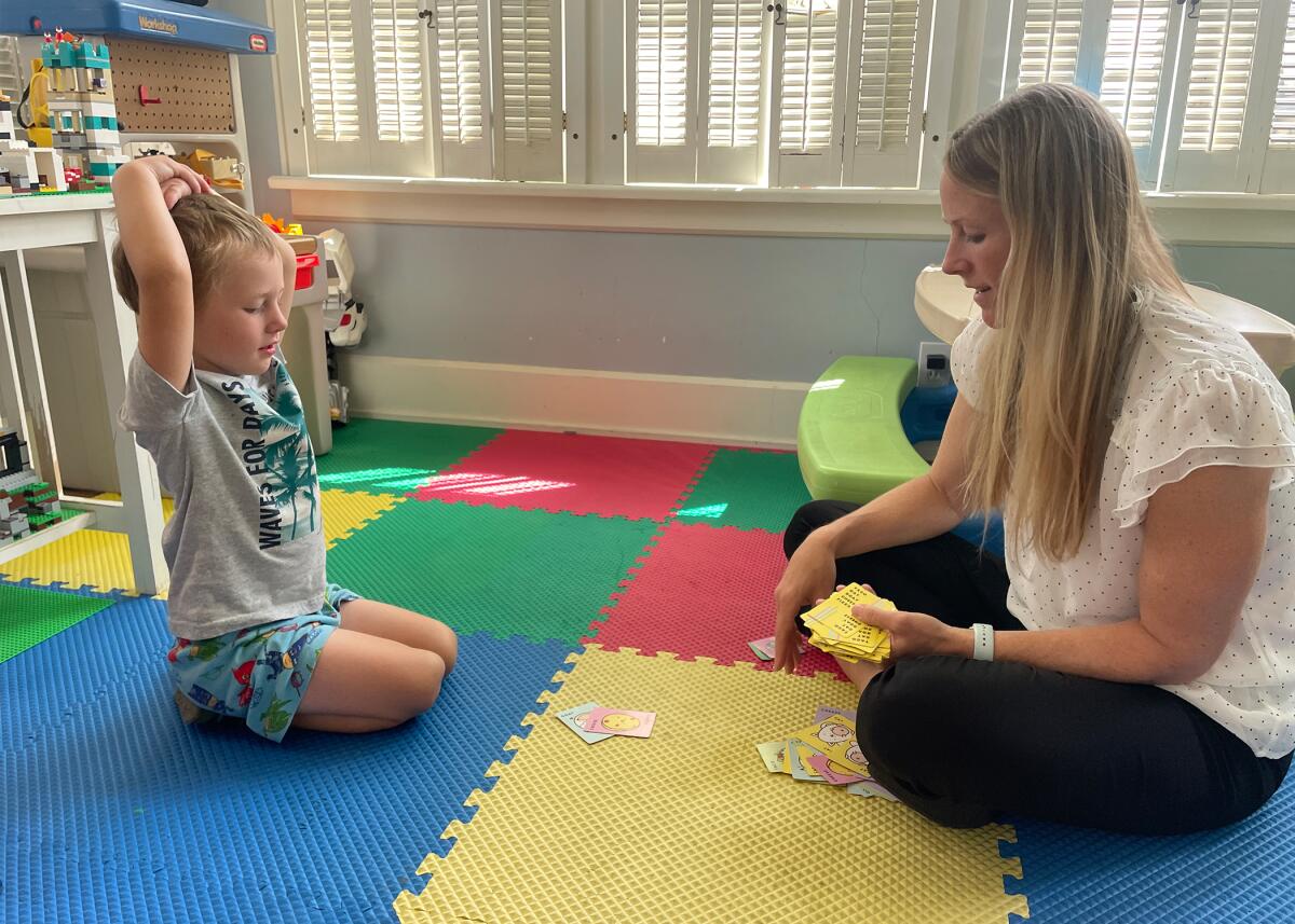 A woman with long brown hair, in white top and dark pants, plays cards with a child while seated on colorful rubber mats