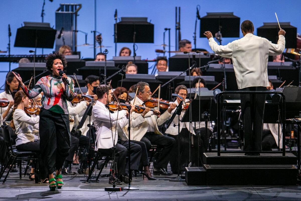Julia Bullock, in a colorful shirt and dark skirt, sings into a microphone while backed by a string orchestra
