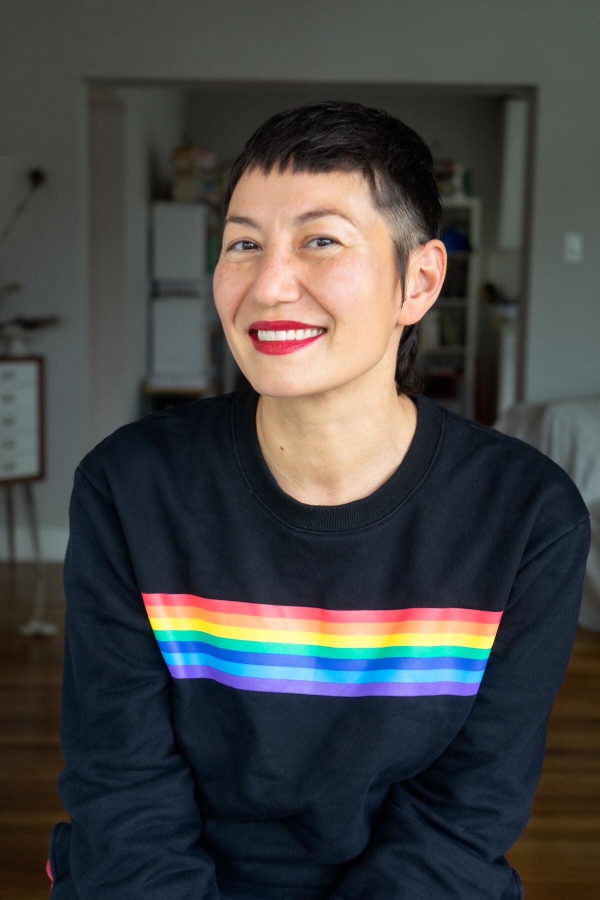 A smiling person wears a black sweatshirt with a rainbow bar across the front of it.