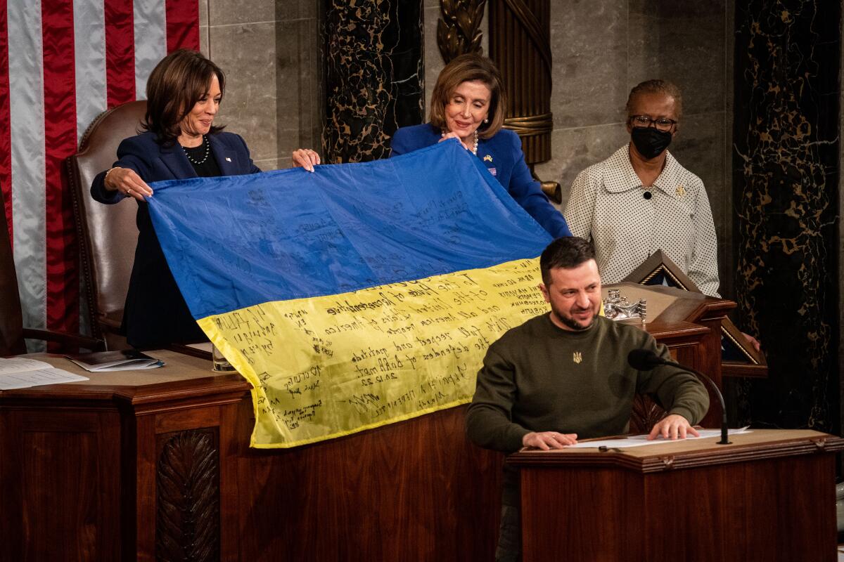 Two women in suits hold a blue and yellow flag behind a man in olive fatigues.