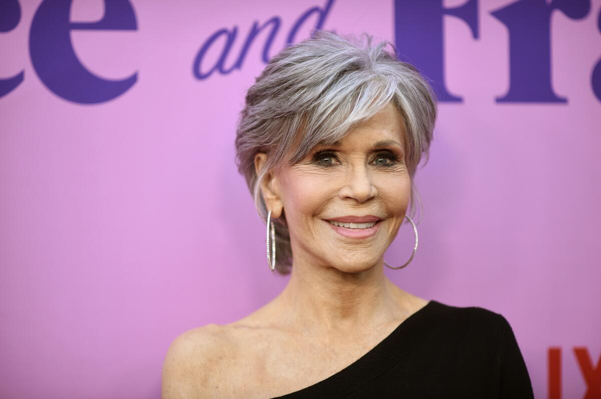 A woman with short gray hair and hoop earrings posing in a one-shoulder black dress