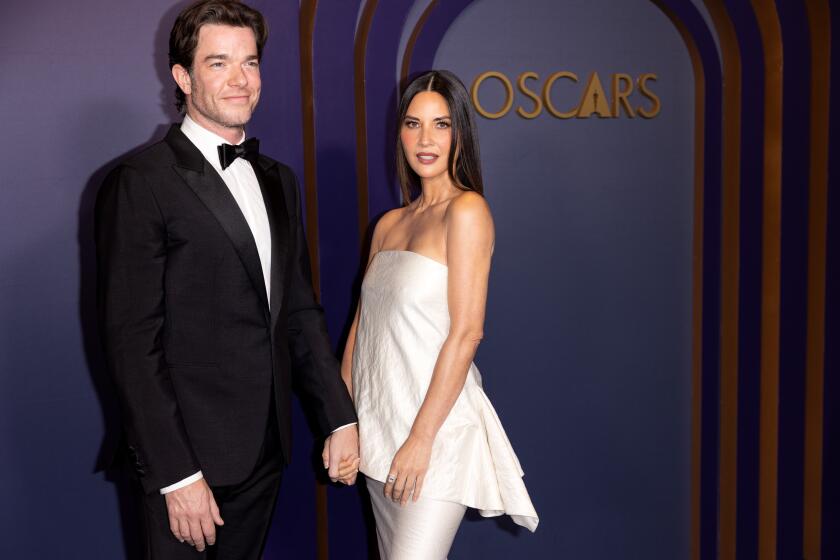 John Mulaney in a tuxedo holding hands and posing with Olivia Munn in a strapless white gown with a peplum at the Oscars