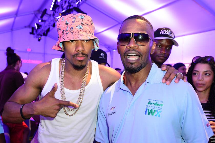 Nick Cannon is wearing a bucket hat and white tank top, posing next to Jamie Foxx who is in a blue polo and sunglasses