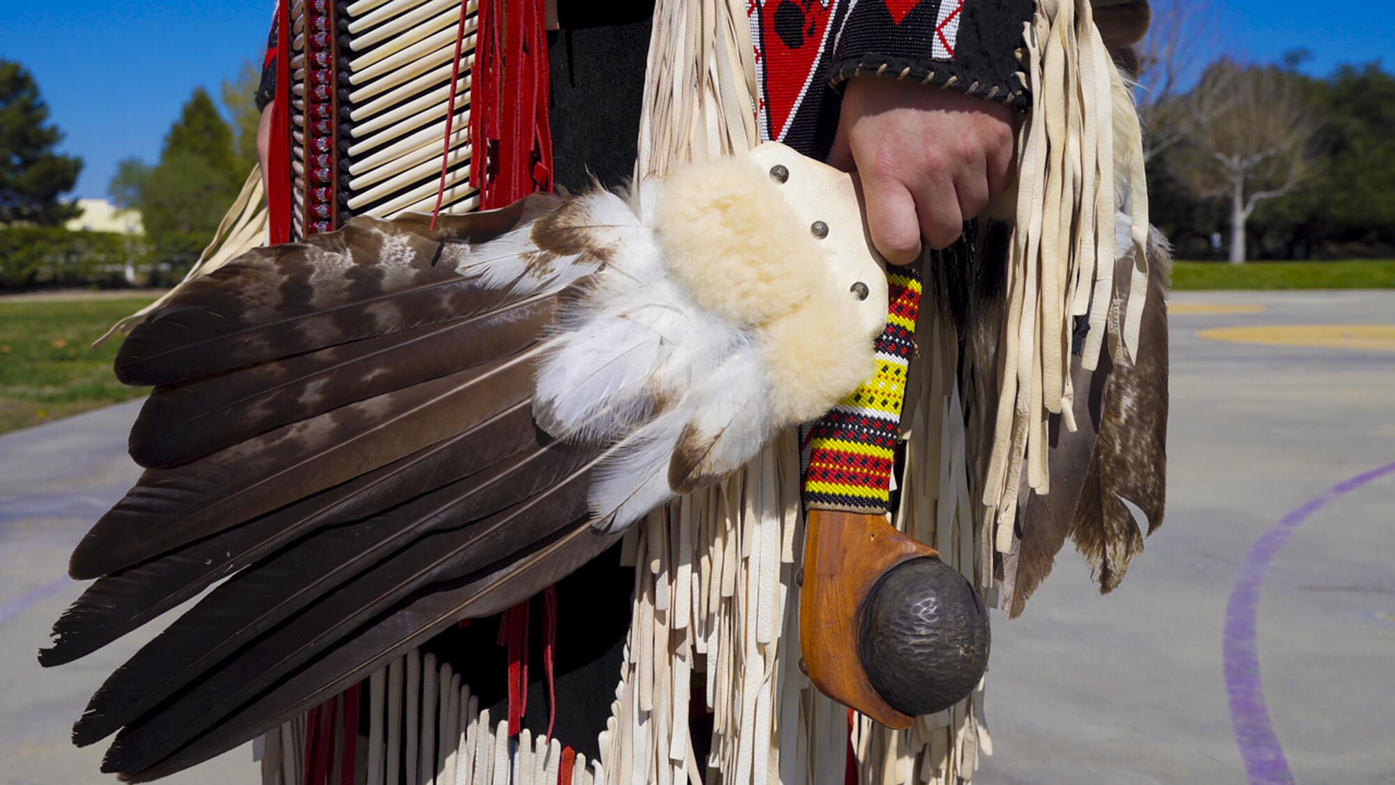 A closeup of a woman's Indigenous fringed dance costume as she holds a handle with feathers in it.