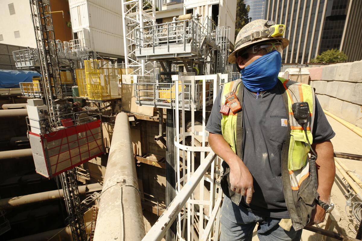 A man with a hard hat and mask stands next to a pit dug for the Metro Purple Line Extension project in Century City in 2021. 