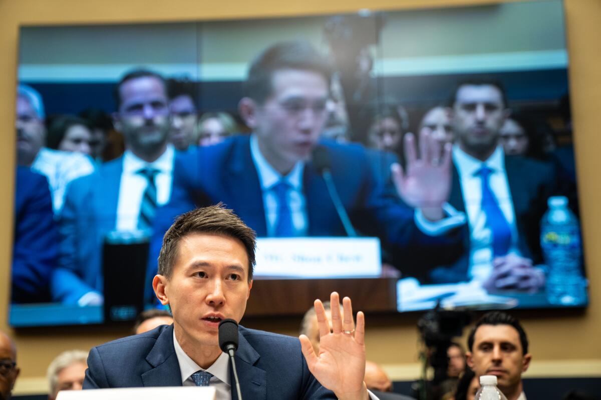 A man with dark hair, in suit and tie, raises one hand while speaking. Behind him is a large screen showing him and others