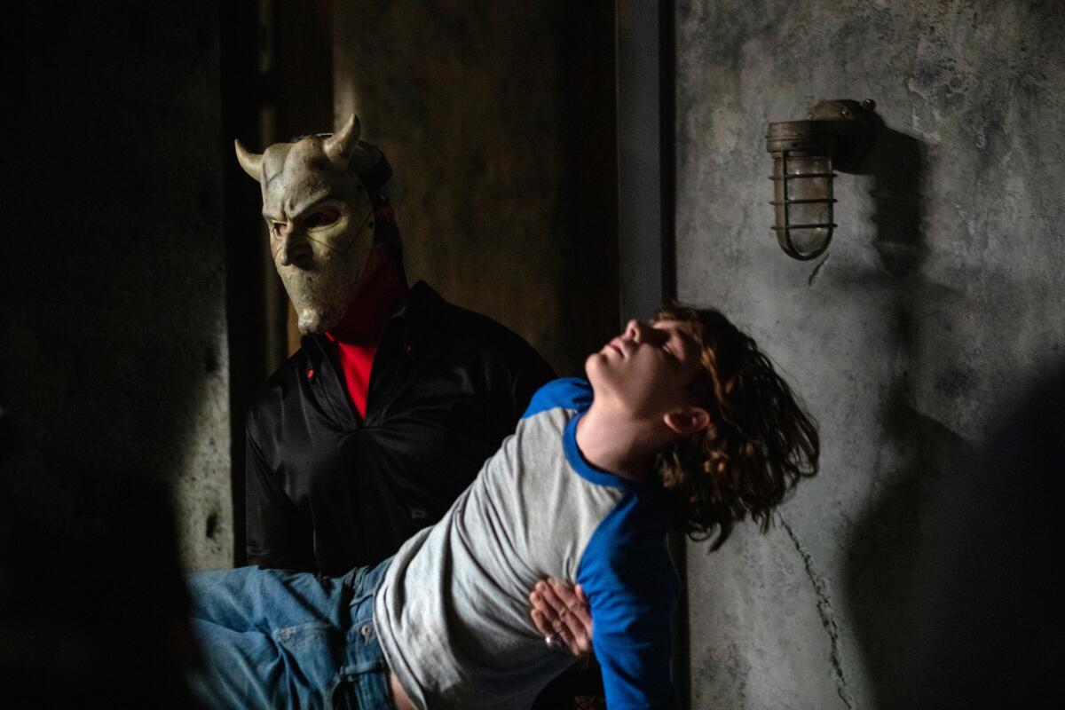 A man wearing a horned mask carries an unconscious boy in the movie “The Black Phone.”