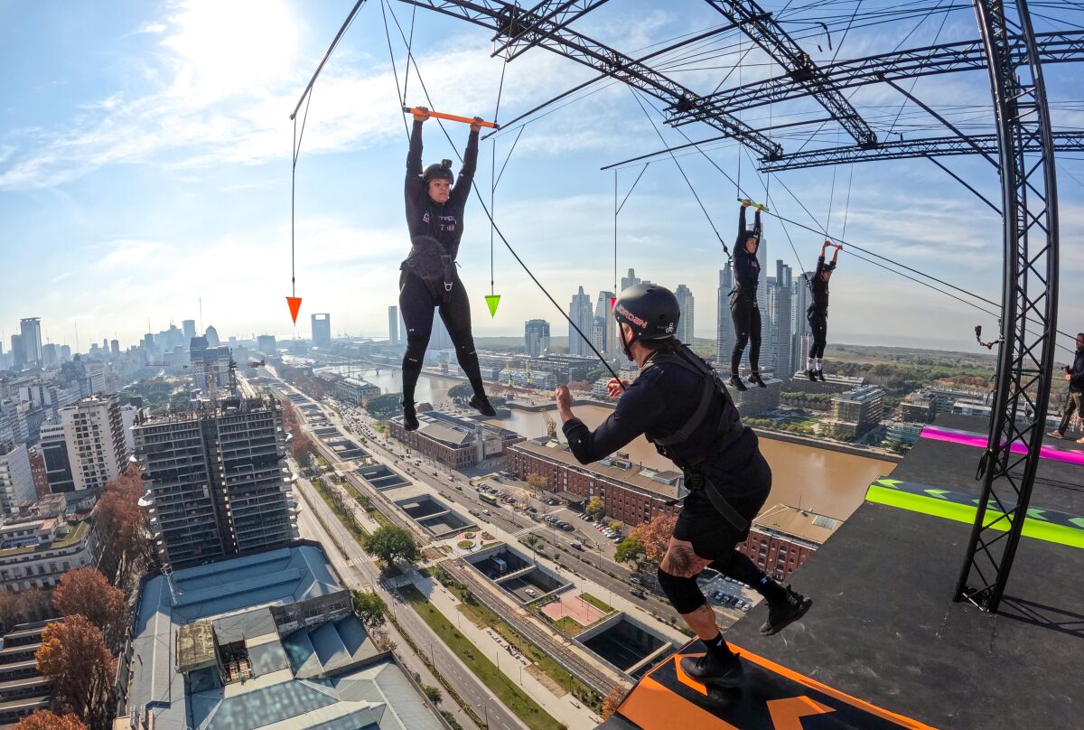 Two people, one hanging from a trapeze, high above a city's skyscrapers.
