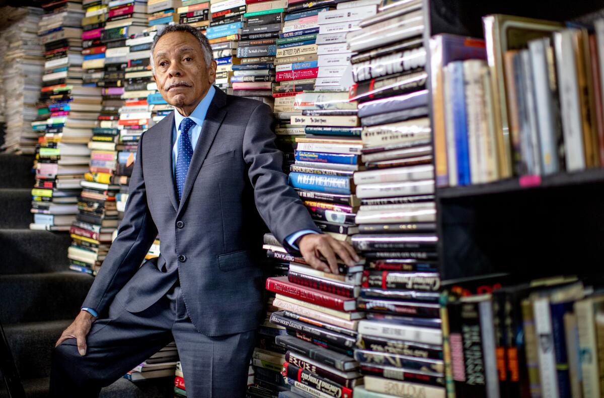 A man in suit and tie stands in front of piles of books.