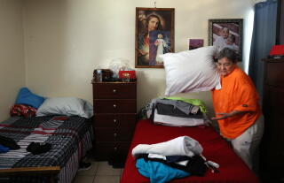 A woman holding a pillow above one of two small beds in a snug room, pictures of Jesus and a photo of two babies on the wall.