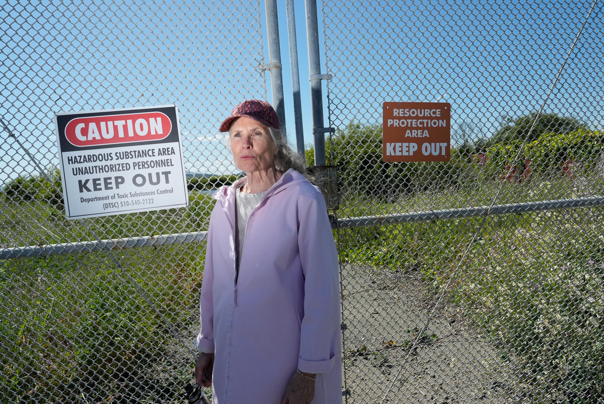 A woman in a purple jacket and a baseball cap stands outside a chain-link fence with "Caution" and "Keep out" signs. 