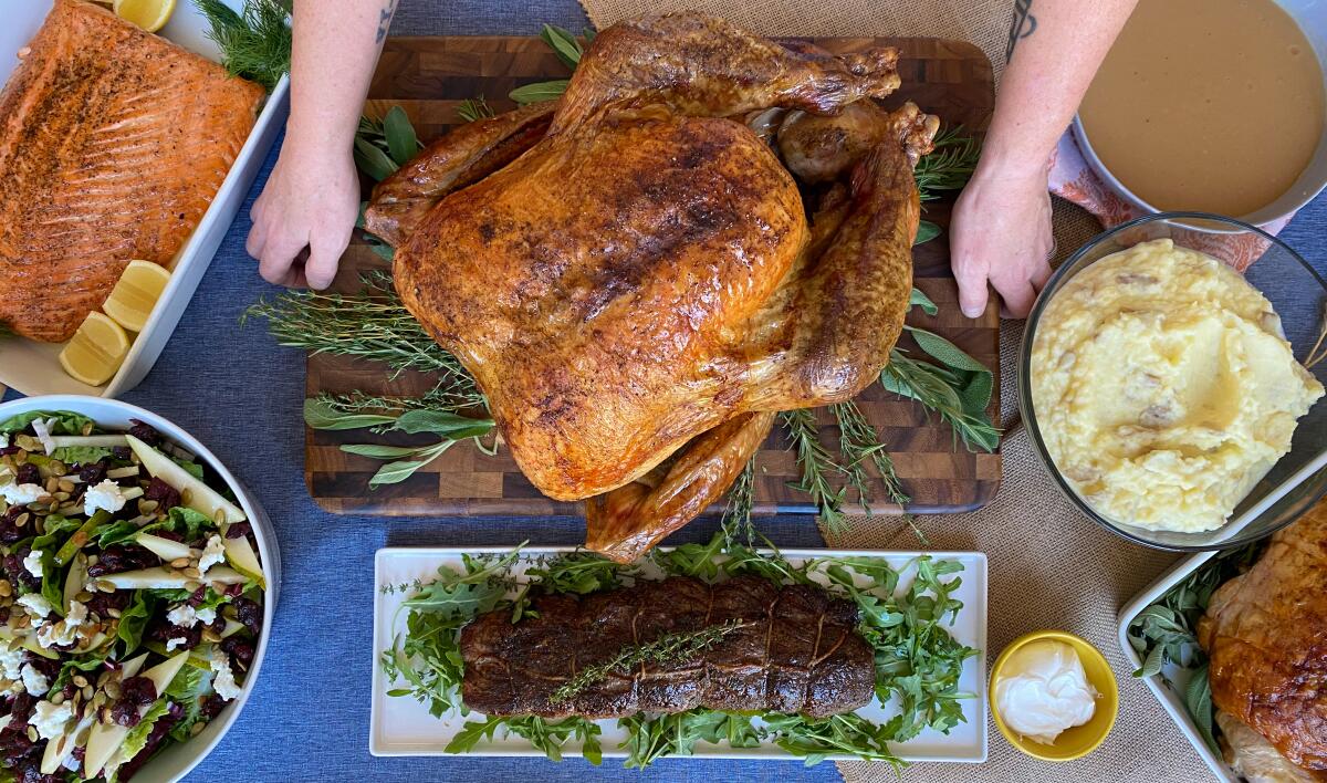 An overhead shot of a roasted turkey on an herb-covered cutting board with salmon and other dishes on a table