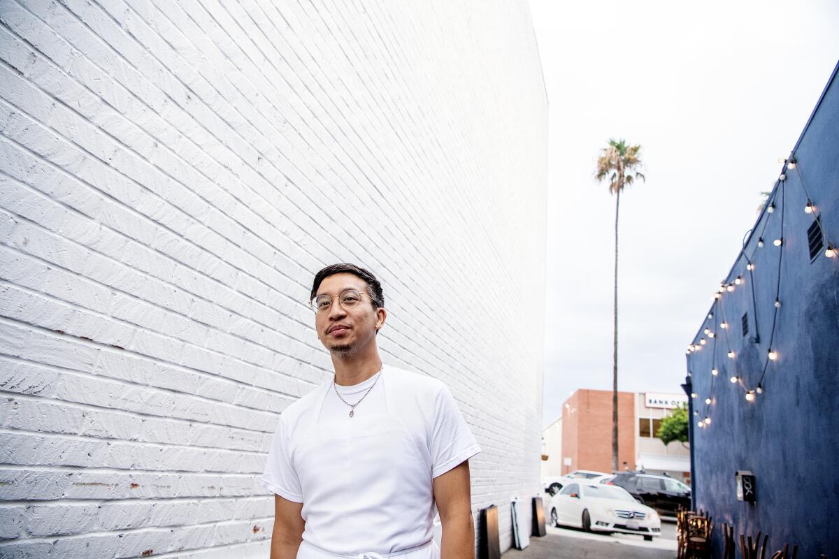 A man in a white T-shirt, glasses and a necklace walks between city buildings; one has a string of lights on it.