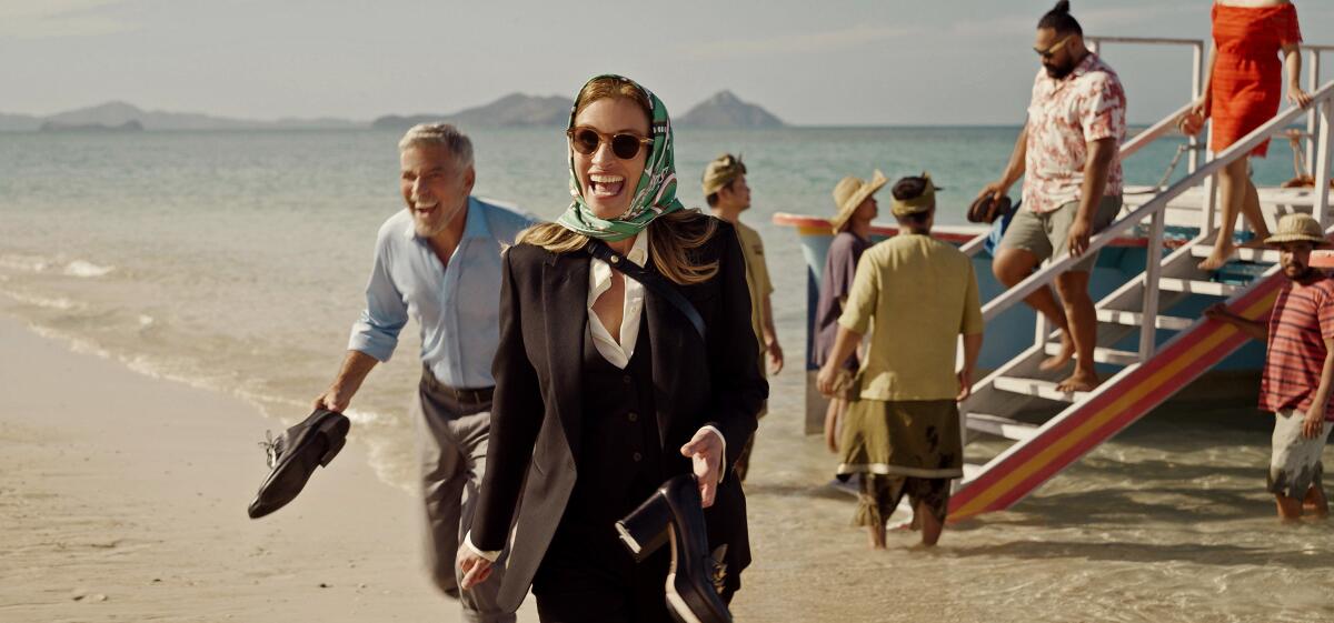 A man and a woman with their shoes in their hands, laughing on a beach in the movie "Ticket to Paradise."