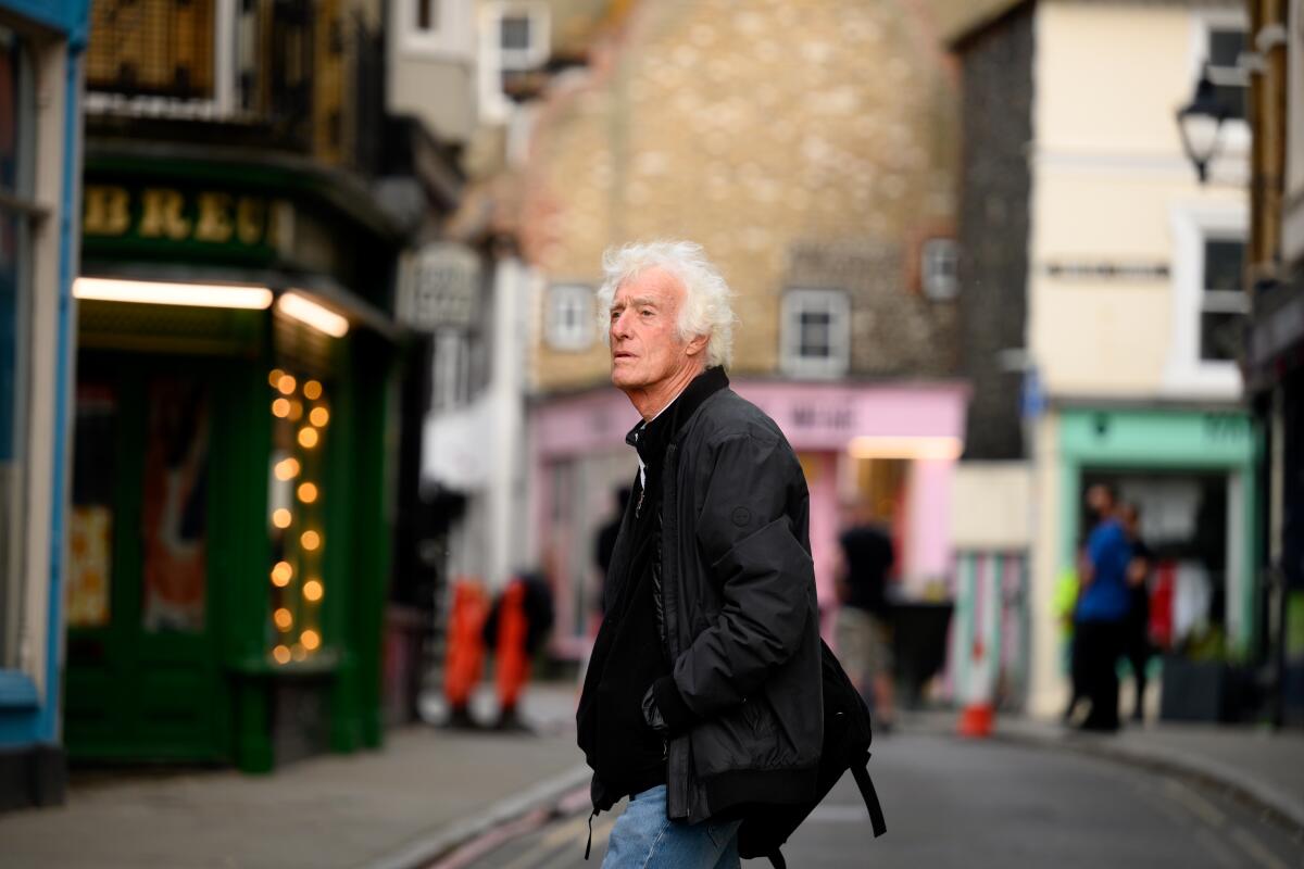 A man with white hair and a dark jacket walks across a street.