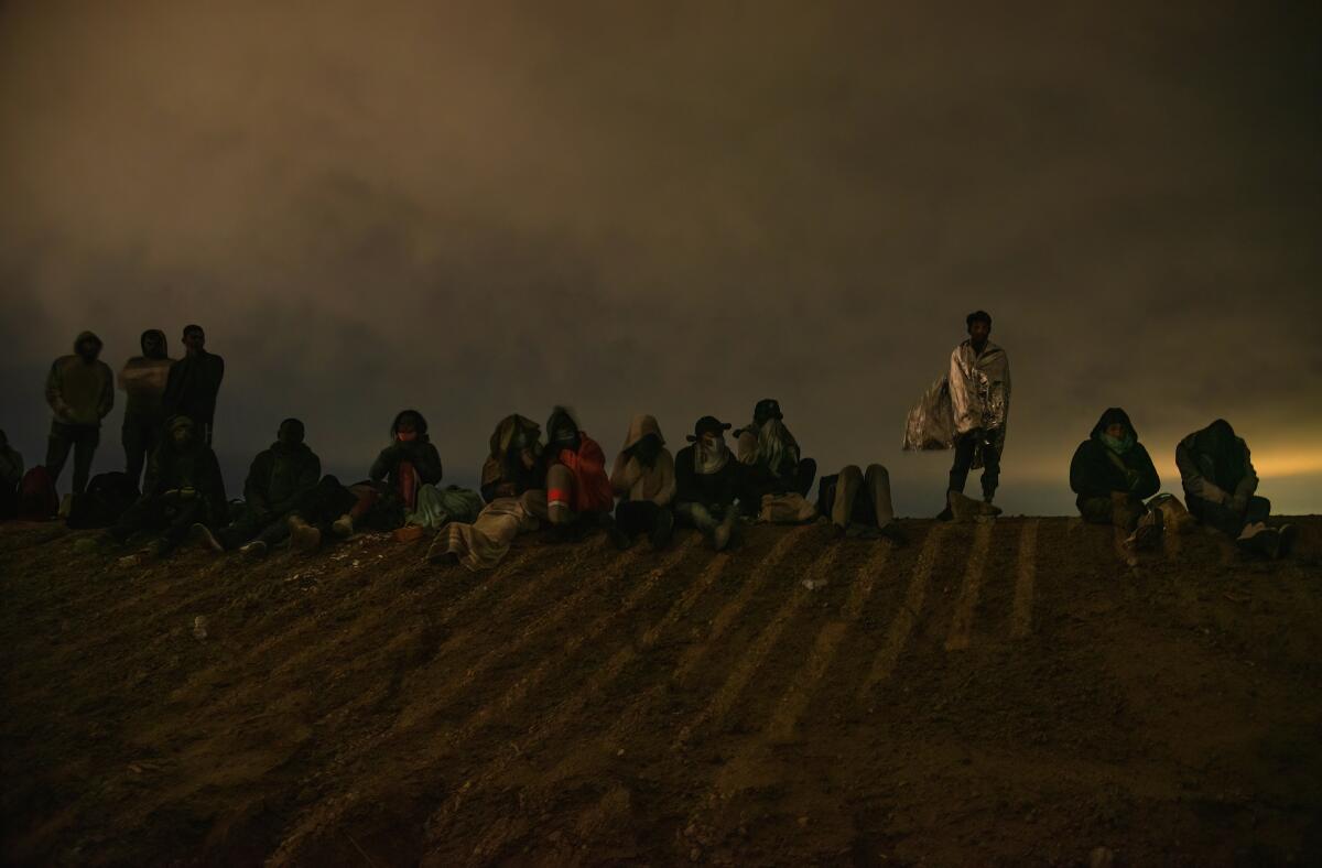 A group of migrants, wearing blankets for warmth, are seen silhouetted against a dark sky. 