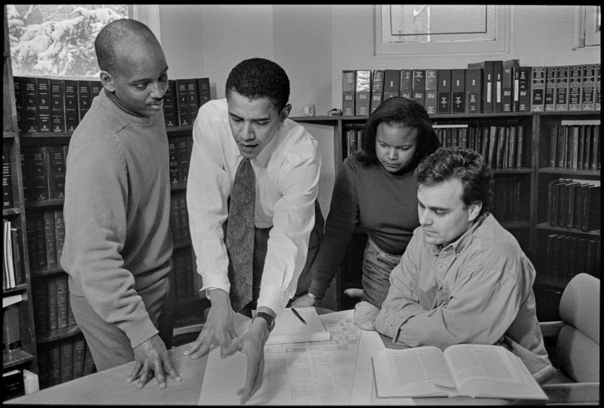 Barack Obama leans over a table in a library, flanked by three people, in a black-and-white image from a 1995 campaign.
