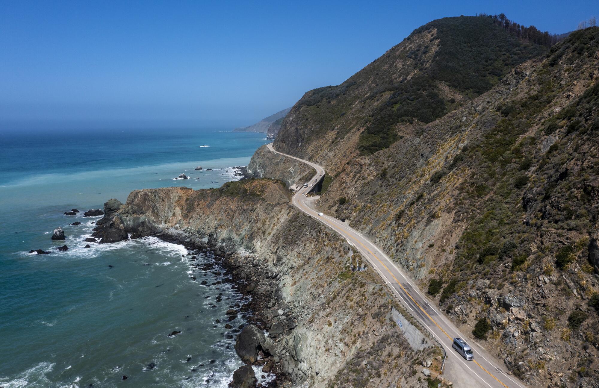 An aerial view shows a two-lane roadway between steep hills and an expanse of ocean.
