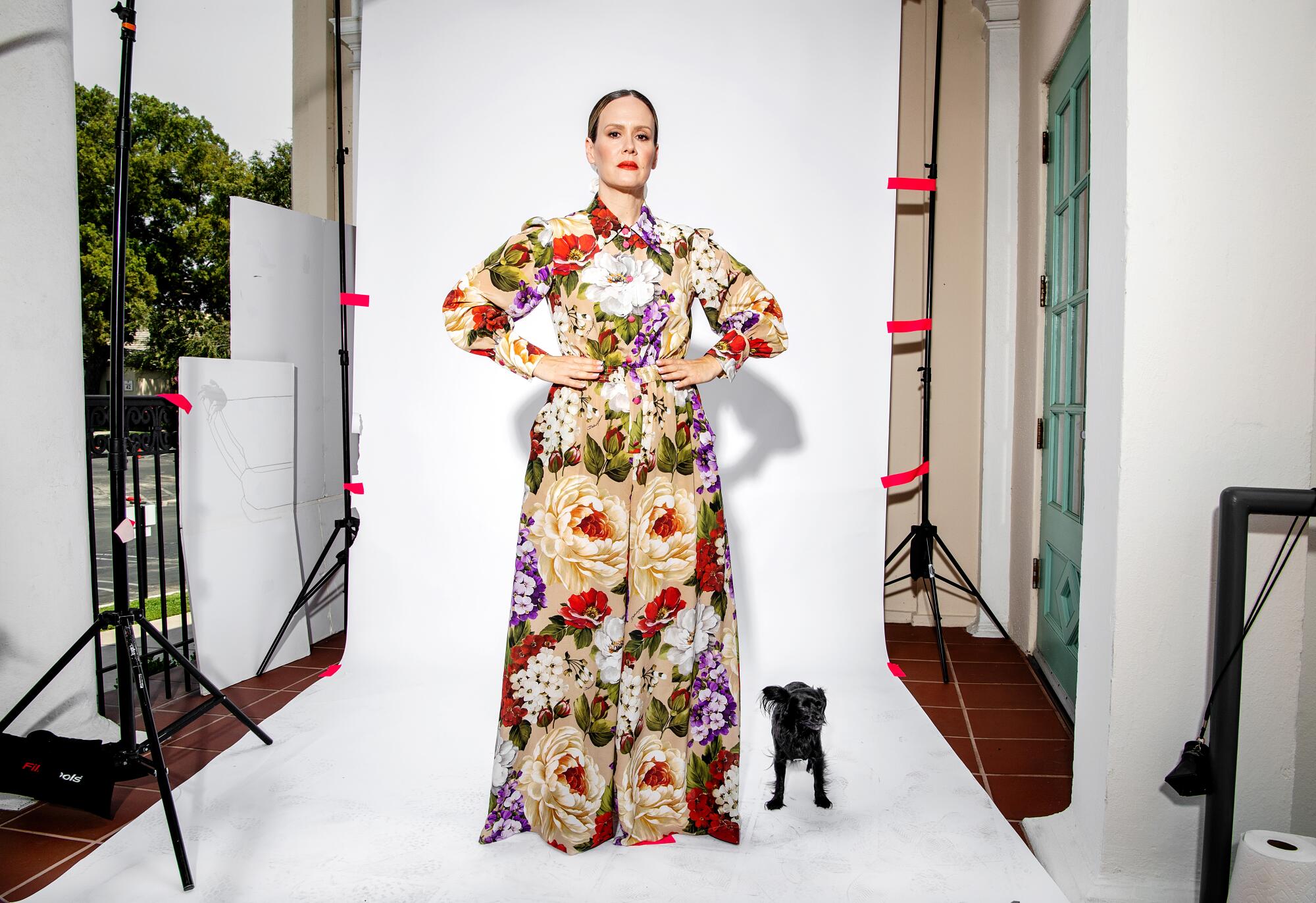 Sarah Paulson stands wearing a colorful floor length flowered outfit with her rescue dog Winnie next to her.