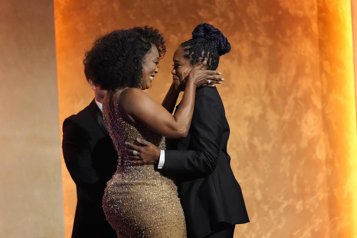 Angela Bassett in a sleeveless gold gown hugs Regina King in a dark suit while onstage at the Governors Awards