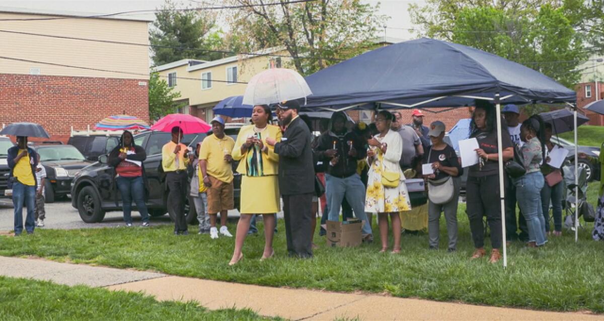 People gather at a prayer vigil under a tent and umbrellas on a rainy day.