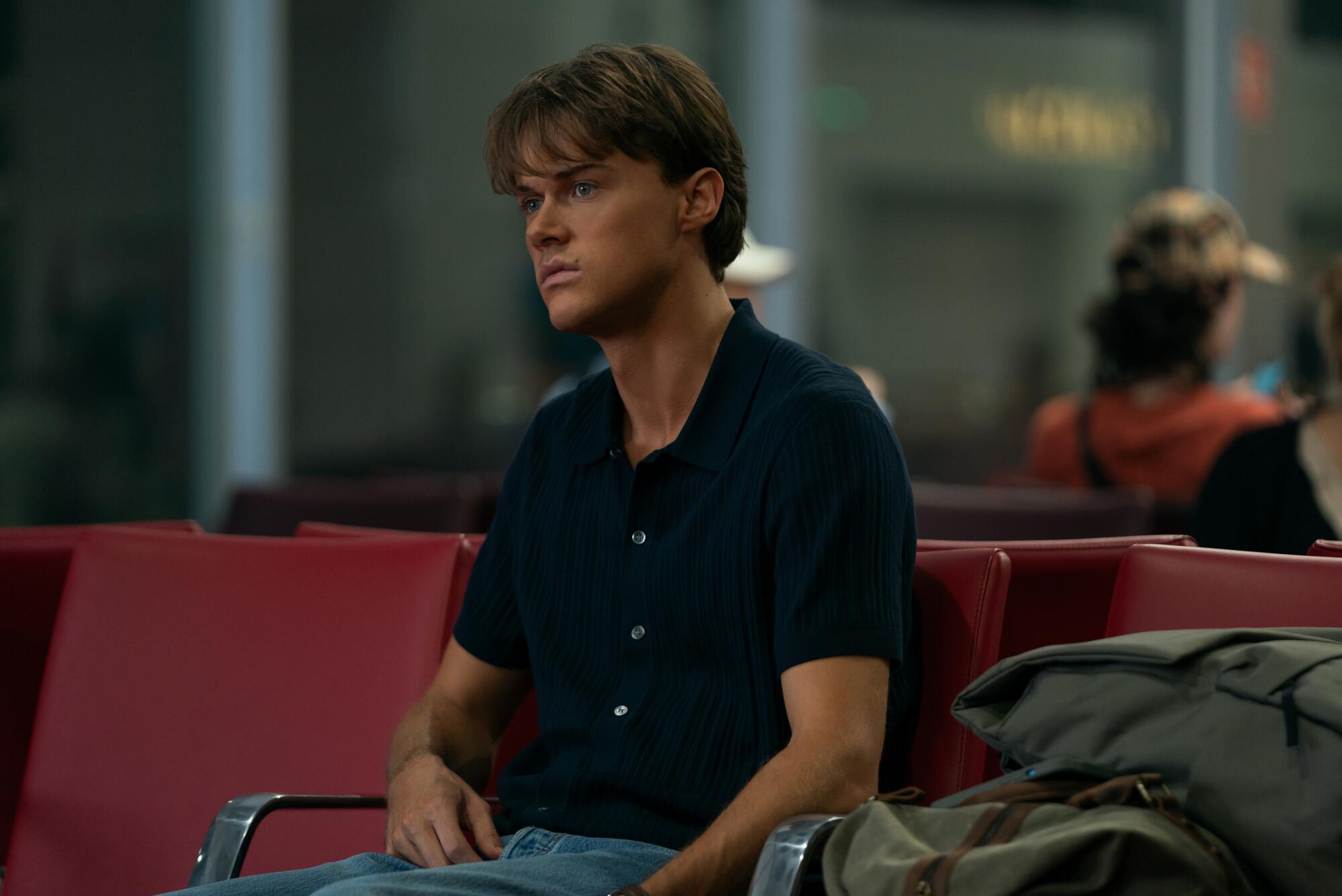 A man in a black shirt and jeans sits in a row of red seats.