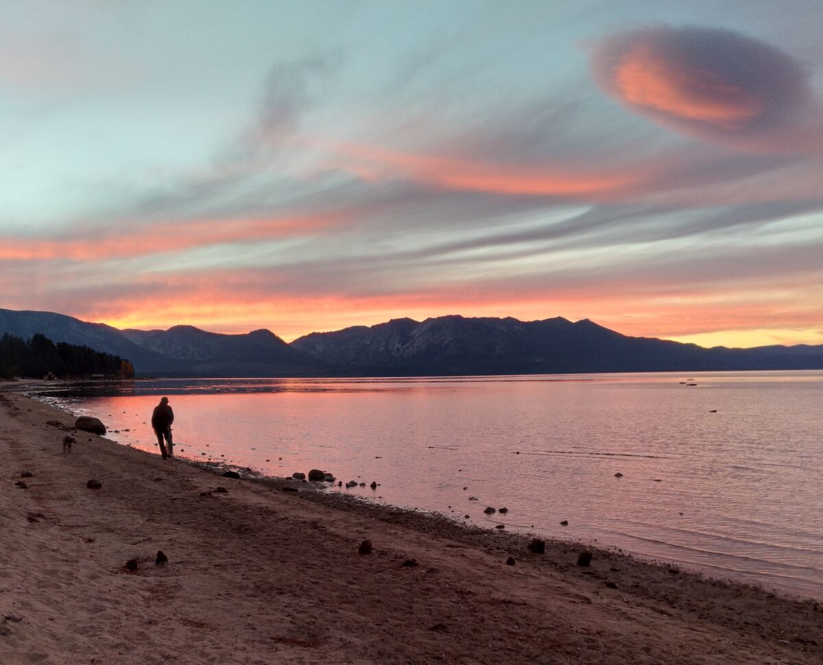 A person walks on the shore of a large lake at sunset with mountains visible in the distance.