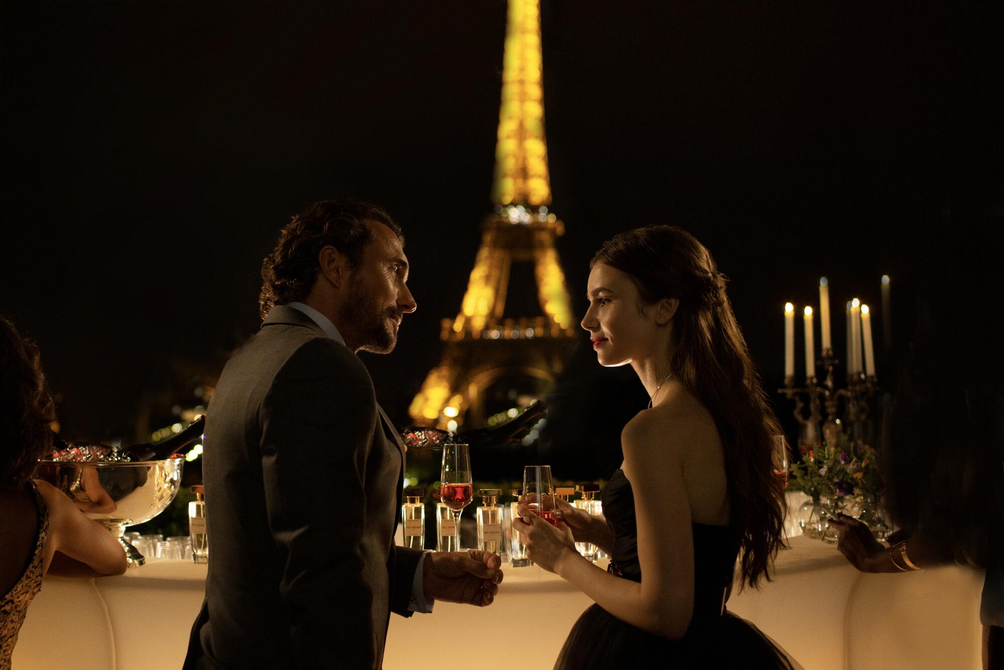 Two actors talk to each other over drinks in front of the Eiffel Tower in a scene from "Emily in Paris"