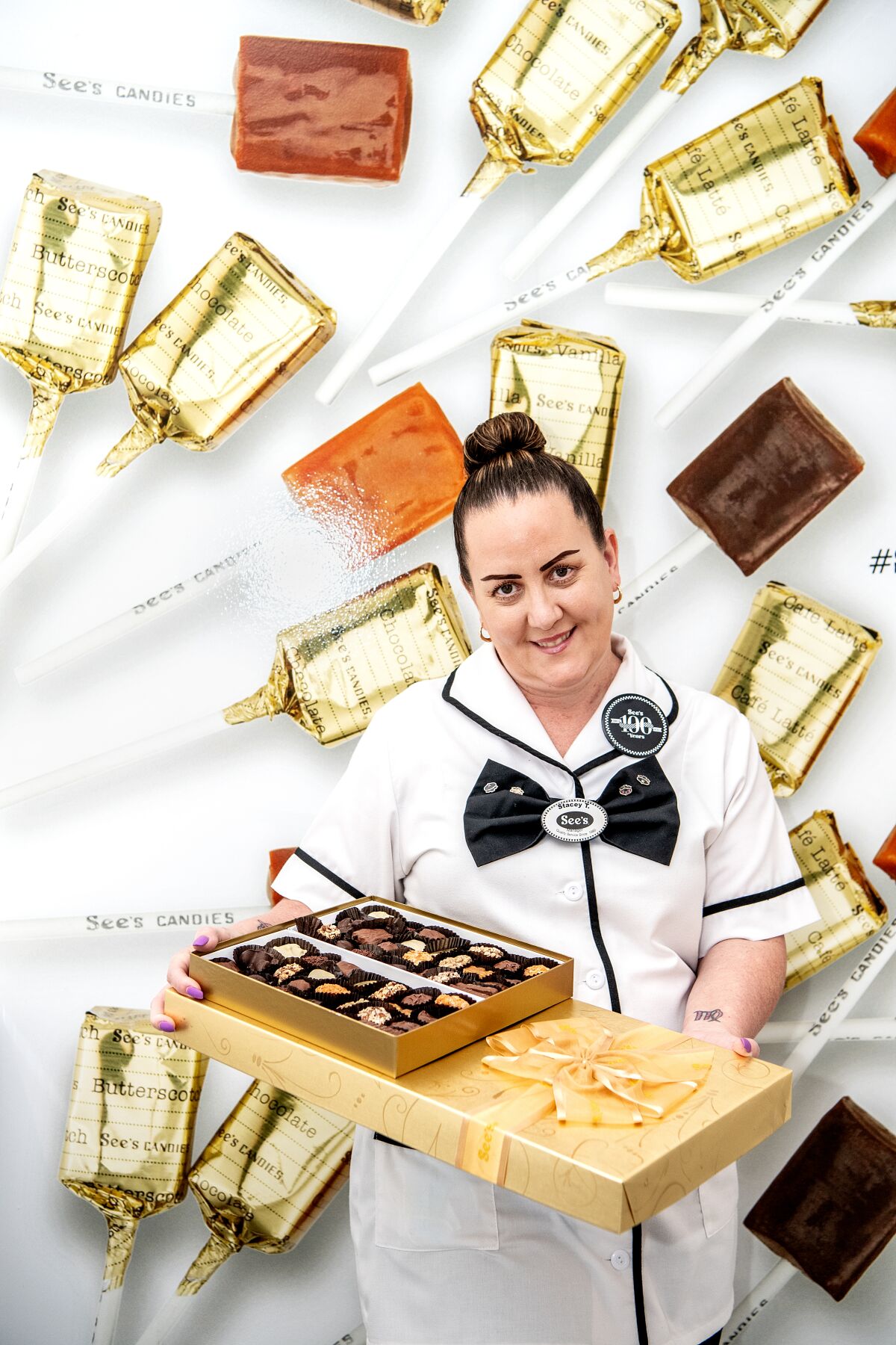 A woman in a white uniform with a black tie holds boxes of See's chocolates.
