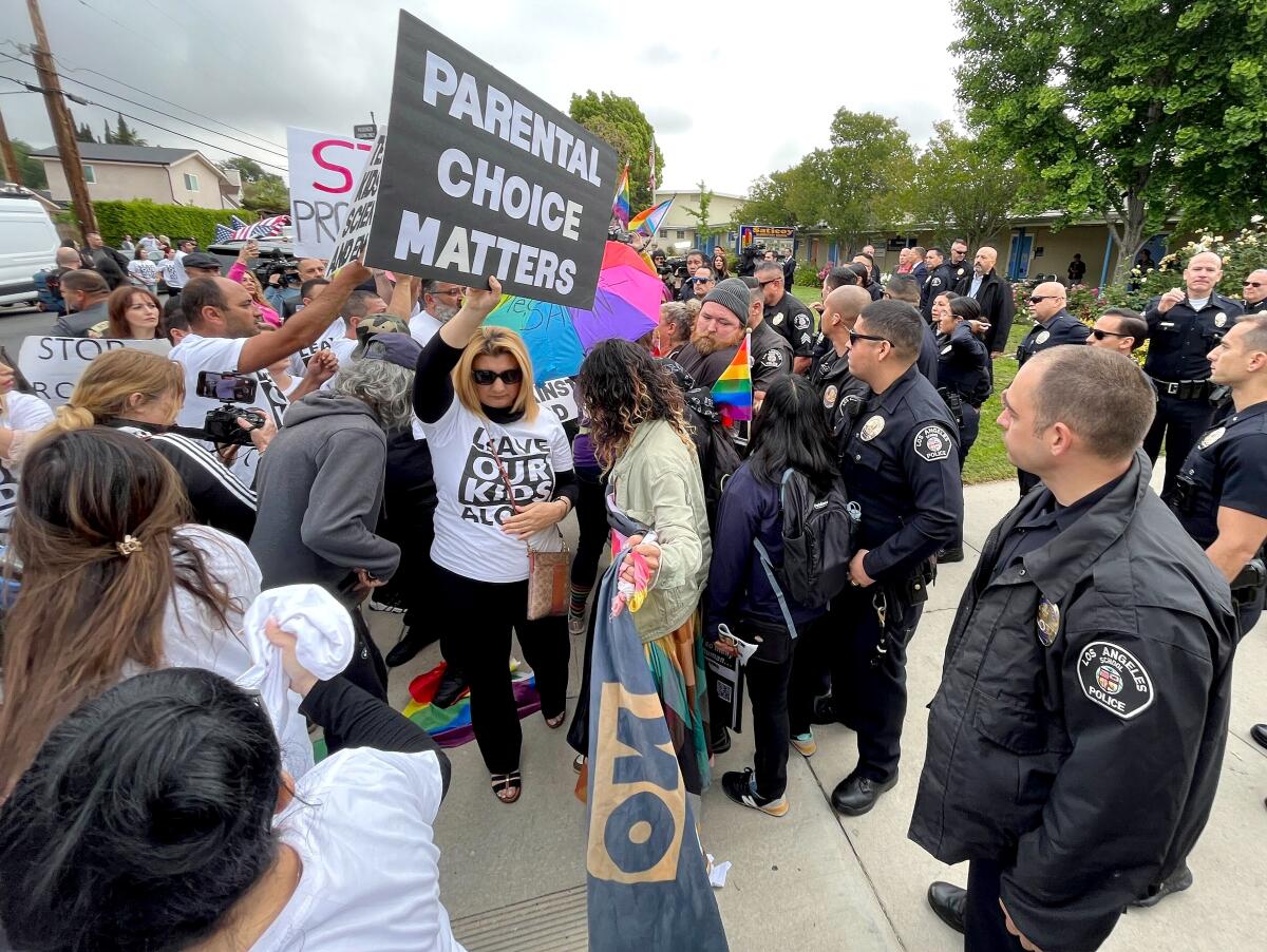 Police officers stand outside a school facing protesters with signs that say "parental choice matters" and other slogans