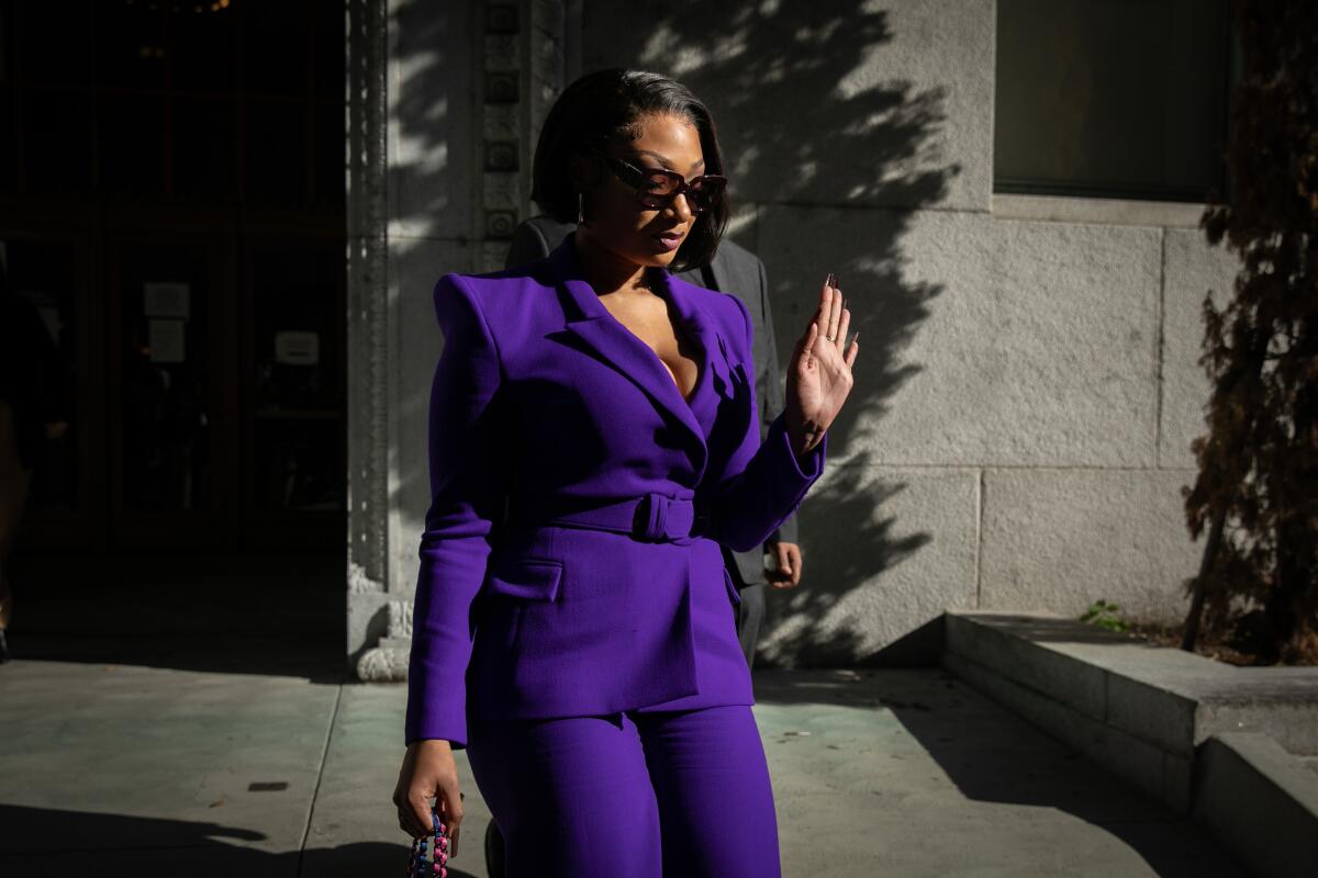 A woman in a purple pantsuit and sunglasses walks outside, holding up her left hand in a "back off" gesture.