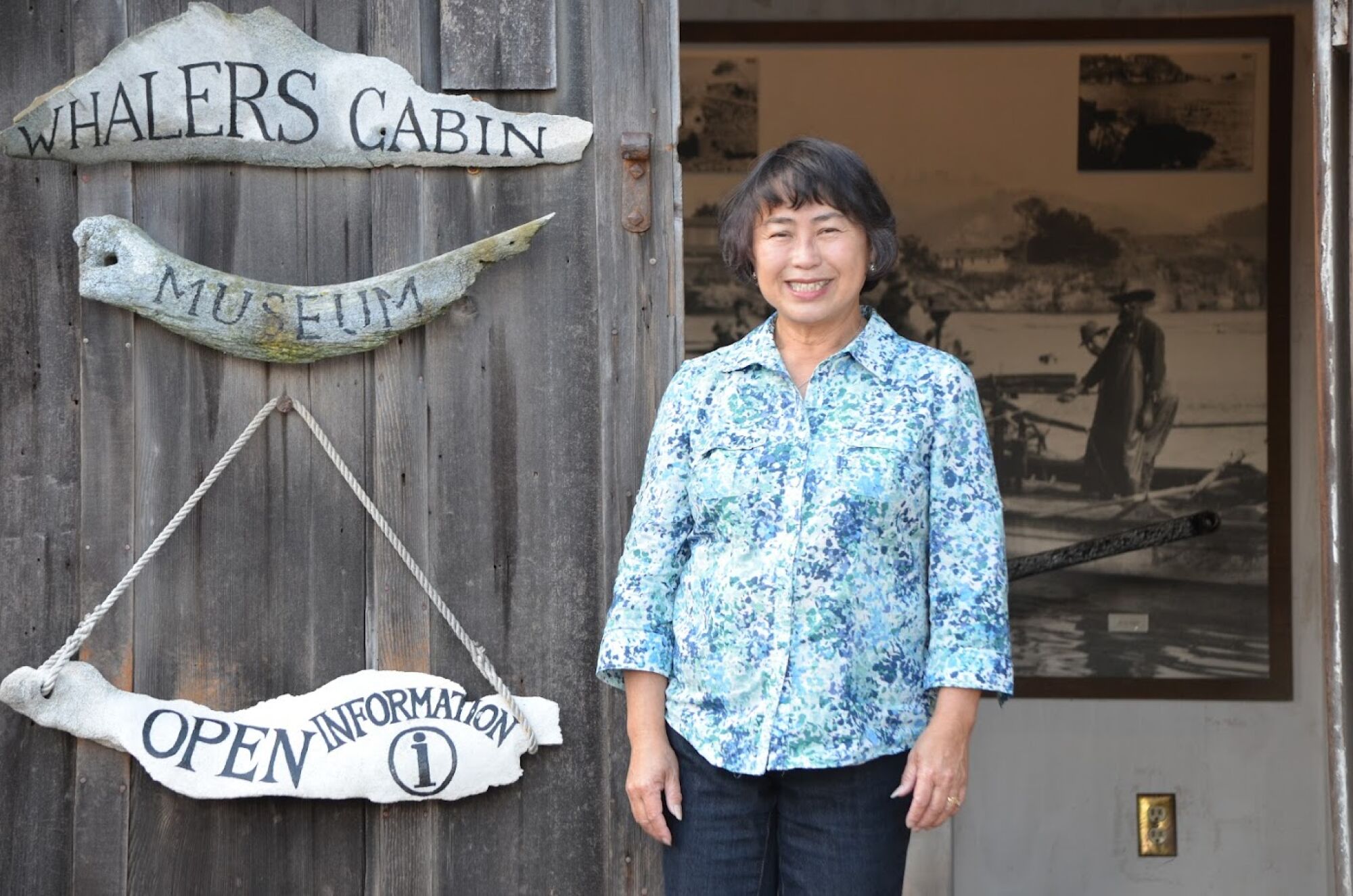 A woman standing in a doorway by signs reading "Whalers Cabin Museum," a large historic photo of a man in a boat behind her.