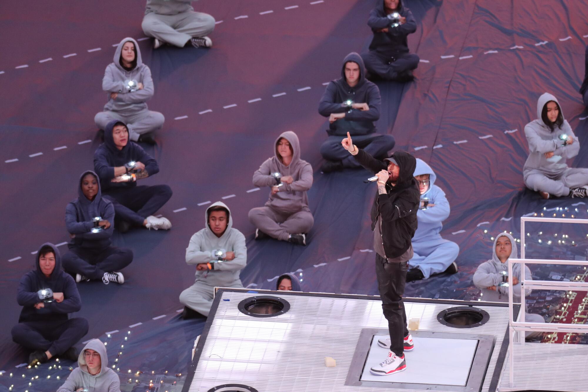 A man in a hoodie onstage, with seated people in hoodies around him. 