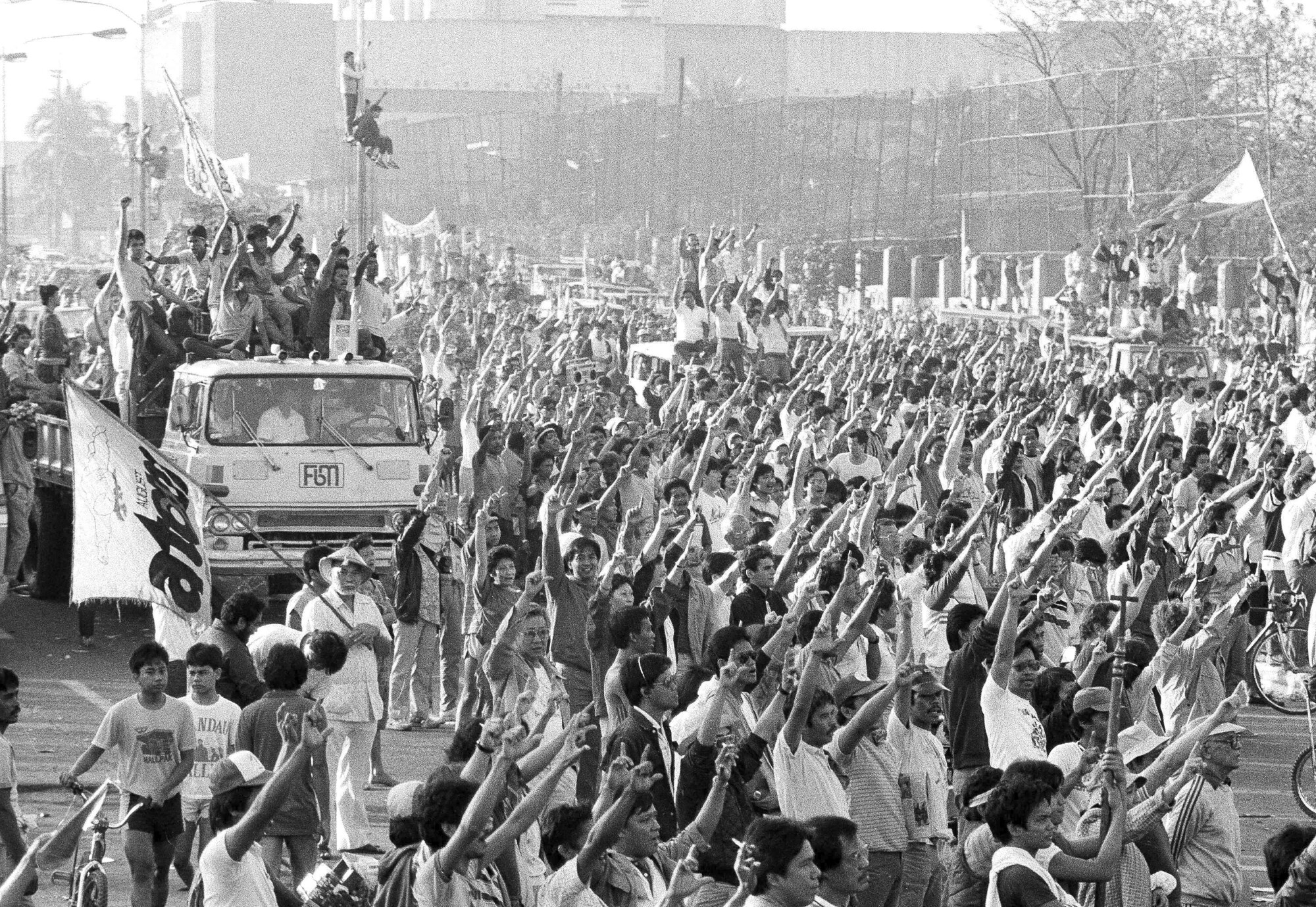 Siyah beyaz bir fotoğrafta, kollarını açmış büyük bir kalabalığın yanında, kollarını kaldırmış insanları taşıyan bir araç görülüyor. 