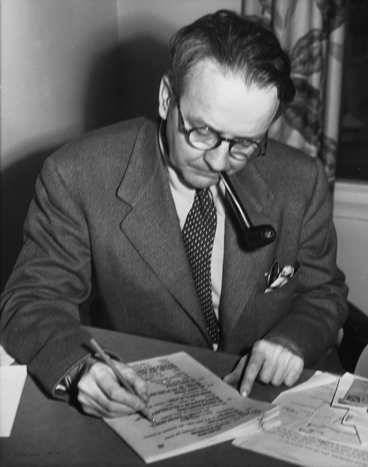 A man in a suit and tie, with a pipe in his mouth, looks down at what he's writing 