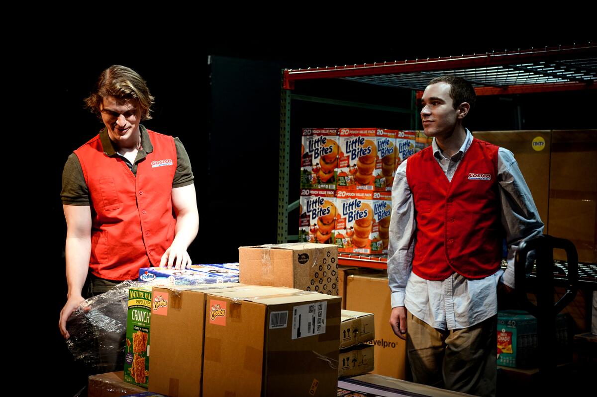 Two actors in red Costco vests onstage amid groceries and boxes in a play