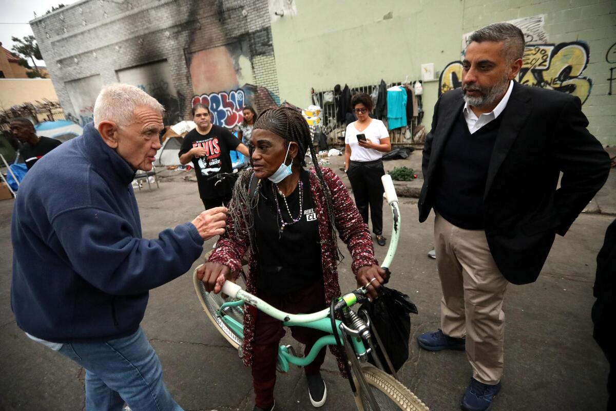 Two men flank a woman on a bicycle on a street near buildings with graffiti on them.